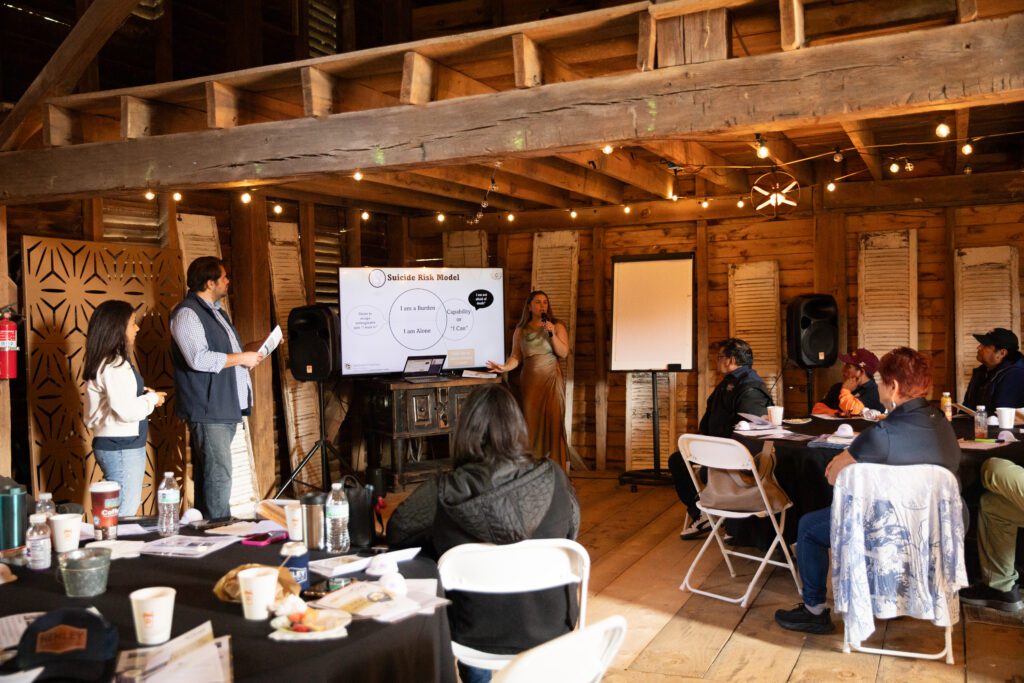 A group of people are attending a Vital Cog Training session in a rustic room. Two presenters stand by a screen displaying a model diagram, while the audience sits at tables with papers and cups.