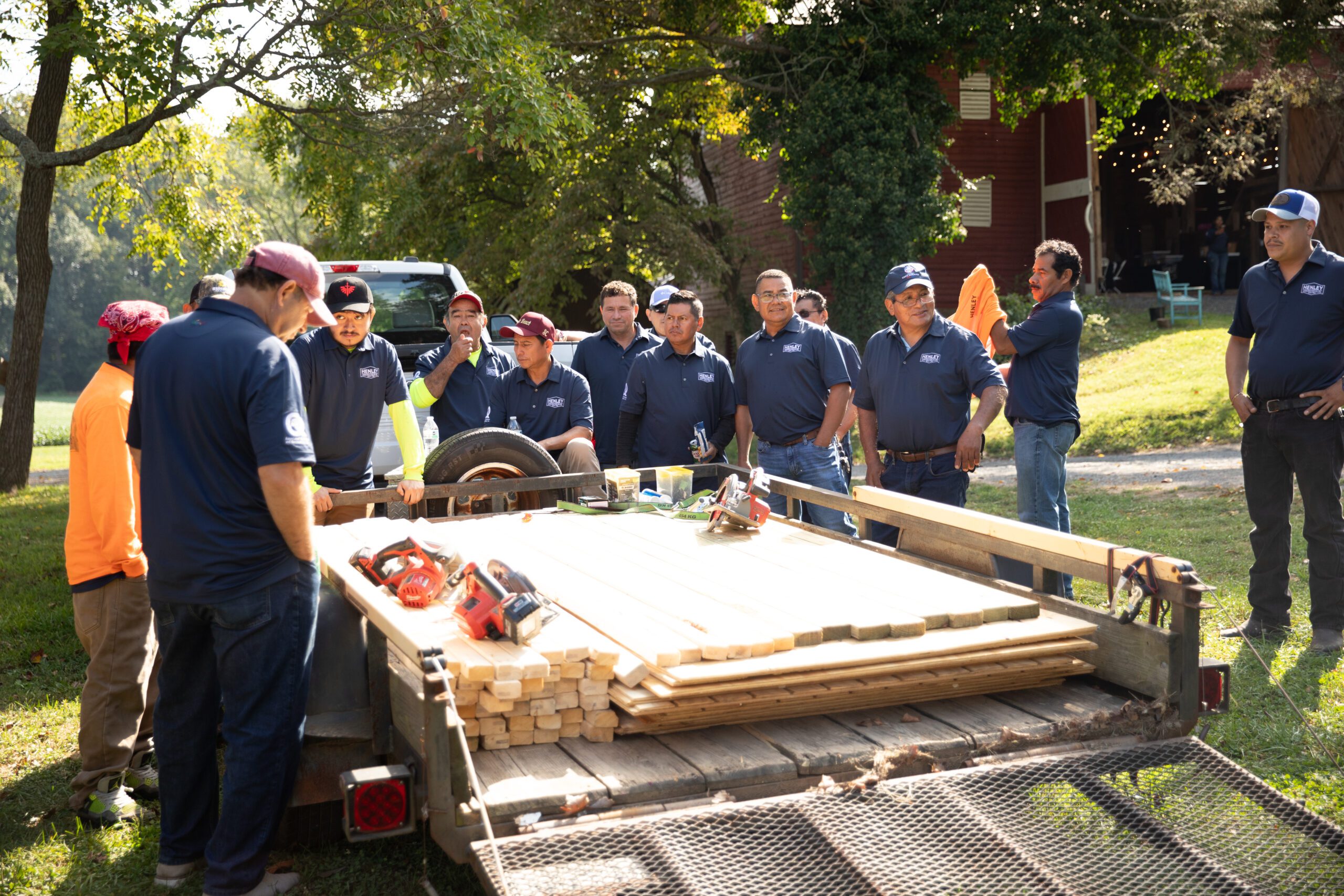 A group of men in blue shirts stand around a trailer loaded with wooden planks and power tools, engaged in discussion outdoors on a sunny day during the Henley Construction Company Summit.