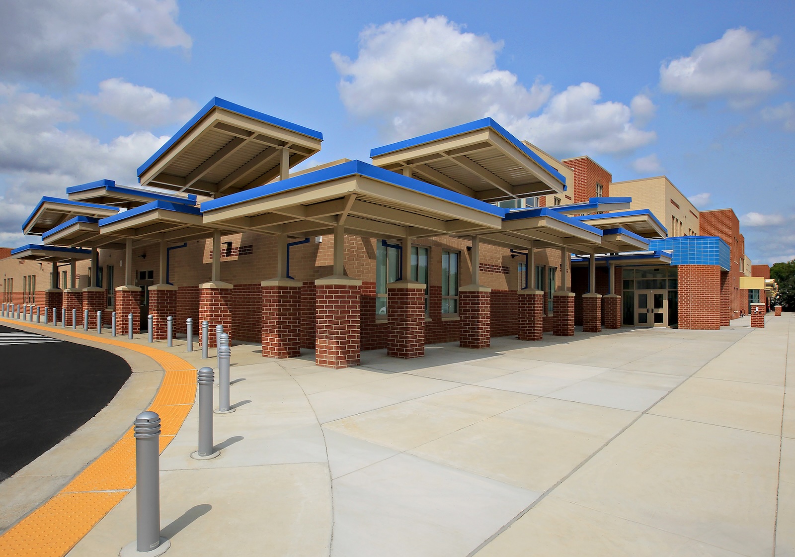 Contemporary building with brick and beige walls, blue trim, multiple canopies, sidewalk, and bollards along the entrance. Clear skies with scattered clouds in the background. This modern design reflects the vibrant atmosphere of Maryvale Elementary School.