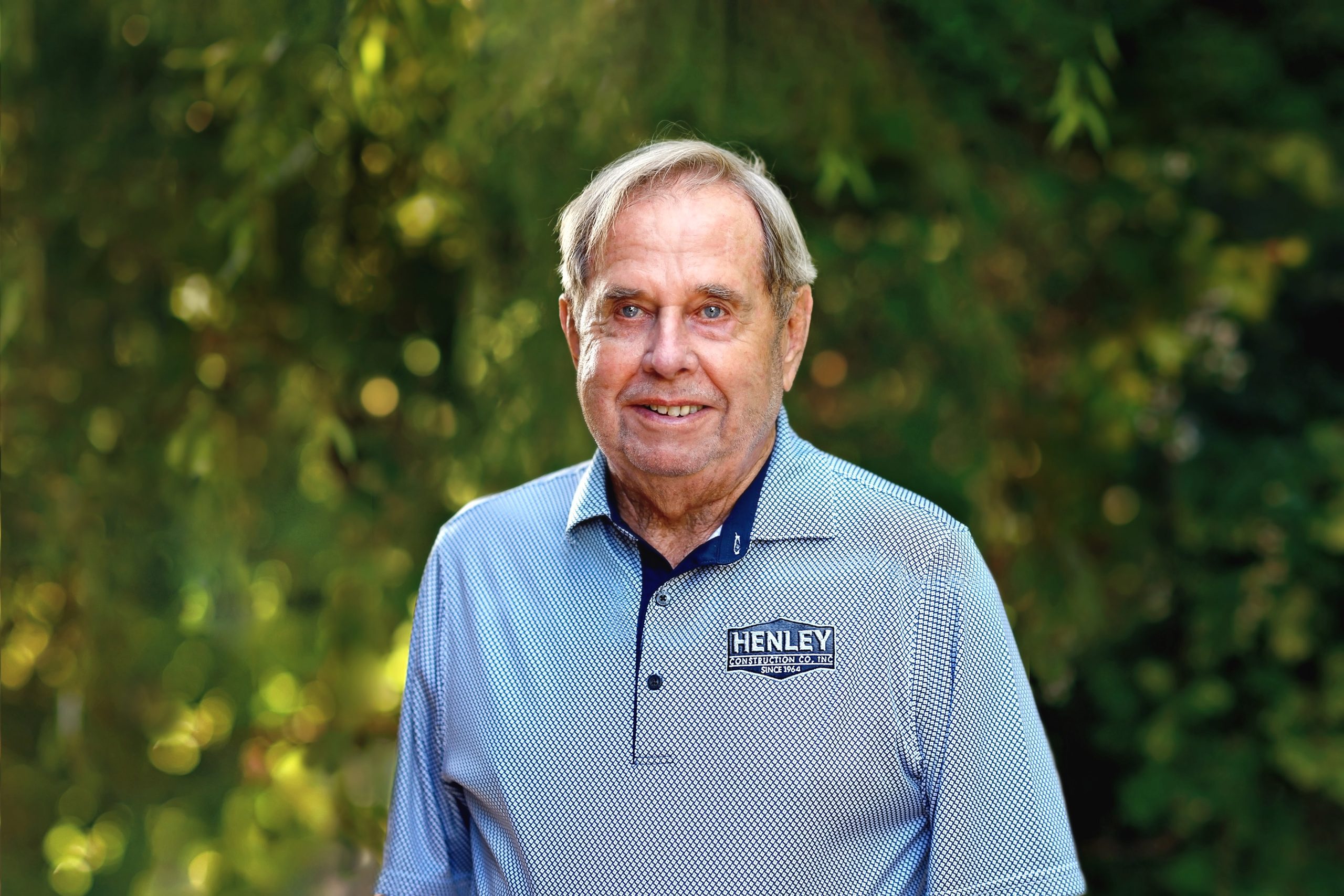 Bob Henley's legacy, stands proudly in a Henley golf shirt against a blurred leafy background, celebrating 60 years of excellence.